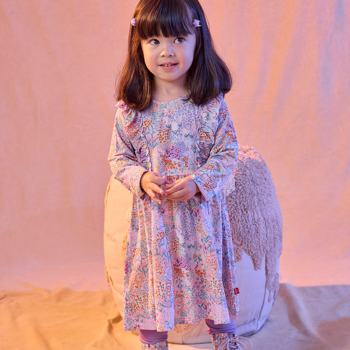 A young girl with dark brown hair wearing a long-sleeved, knee-length floral dress in pastel colors, standing in front of a draped beige fabric backdrop.