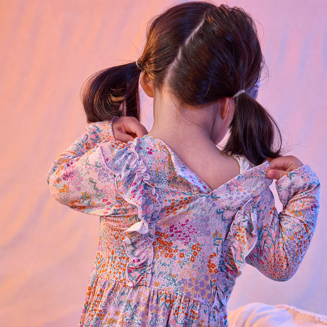 A young girl viewed from behind wearing a long-sleeve floral dress with ruffled details, holding the sides of her back open. She has dark brown hair in two pigtails and is posed against a soft pink fabric backdrop.
